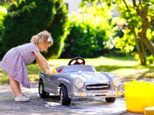 Søt liten jente i sommerkjole, som vasker tråkkebilen sin med vann fra en gul bøtte