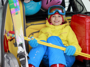 gutt i gult og blått slalomutstyr, sitter i bagasjerommet på en bil klar for påskeferie