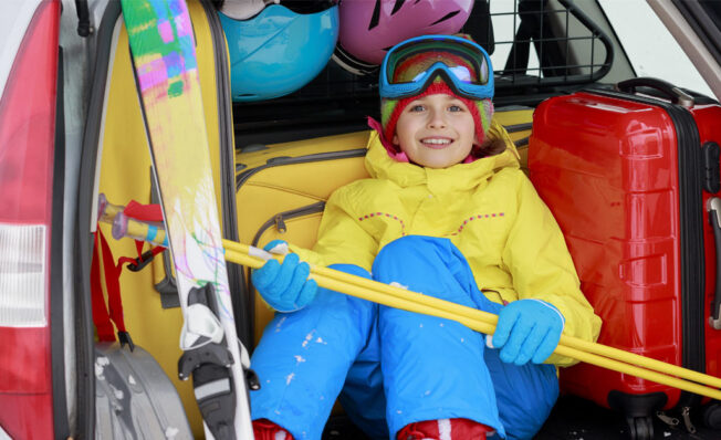 gutt i gult og blått slalomutstyr, sitter i bagasjerommet på en bil klar for påskeferie