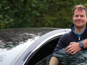 En fornøyd mann står i døren på sin Tesla etter at han byttet til Mekonomen og sparte 40.000 kr på reparasjon etter EU-kontroll.