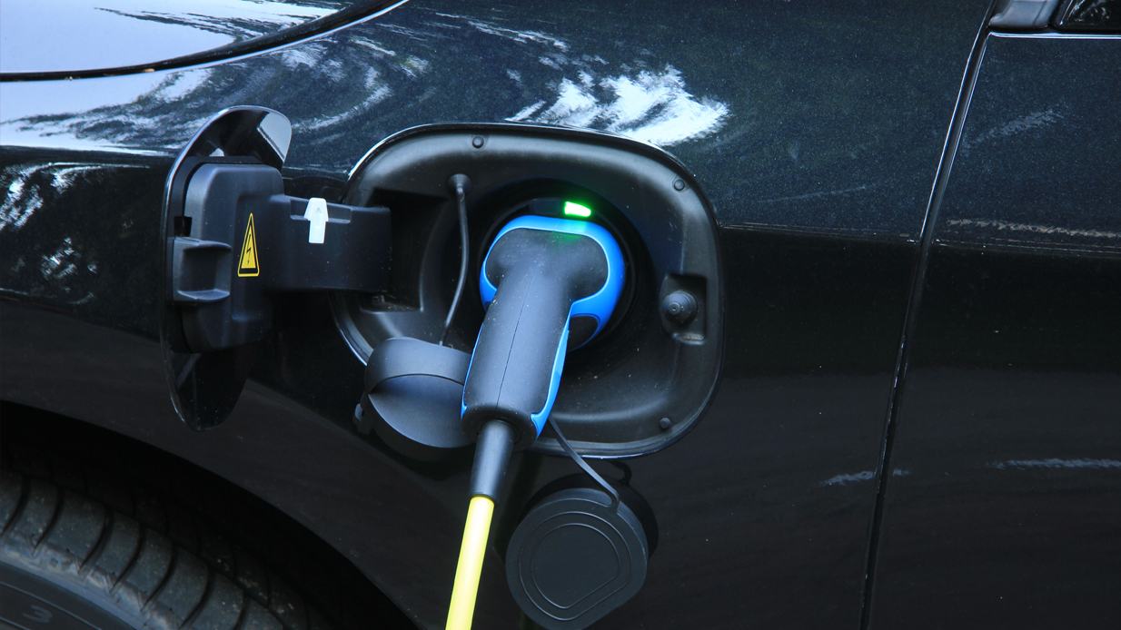 Den blå og sorte Elbilkontakten er plugget inn i ladekontakten på en mørkblå elbil