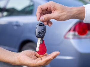 Bilnøkler blir overlevert fra en mannshånd til en dames hånd foran en blågrå bil