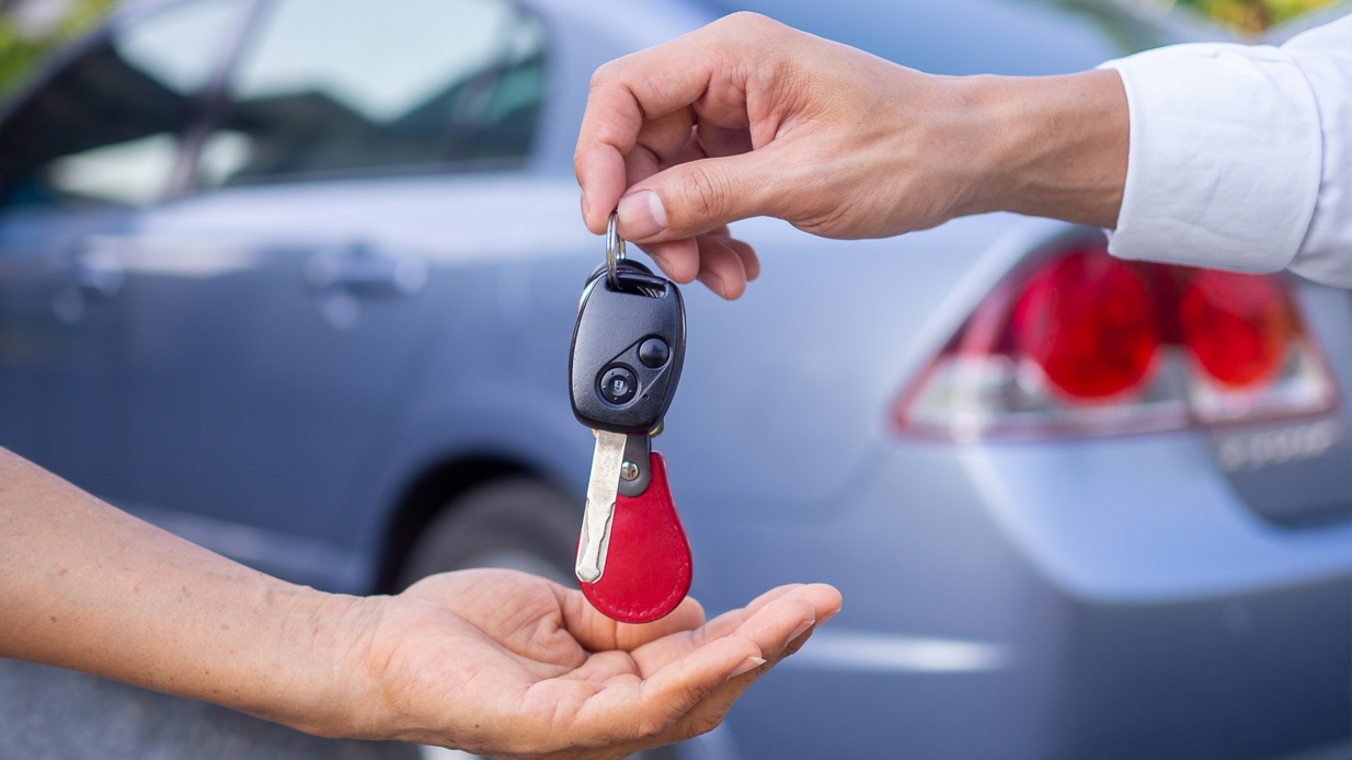 Bilnøkler blir overlevert fra en mannshånd til en dames hånd foran en blågrå bil