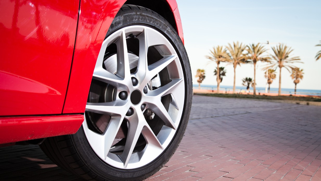 Rød sportsbil parkert på rød brostein med utsikt til hav, strand og palmer – illustrasjon til «Tenk på dette når du kjøper sommerdekk»