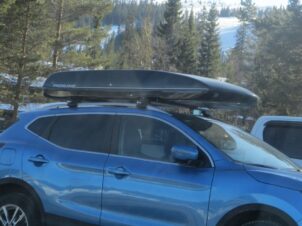 blå bil med skiboks på taket, parkert på en parkeringsplass med skog og skibakker i bakgrunnen