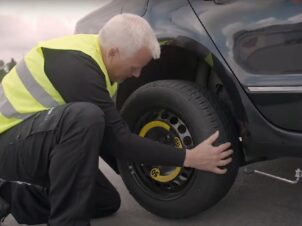 Mann med gul refleksvest skifter hjul på en punktert mørk bil