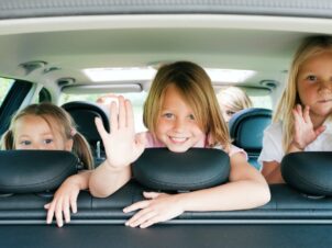Bilferie - 3 glade barn vinker bakover fra baksetet på bilen.